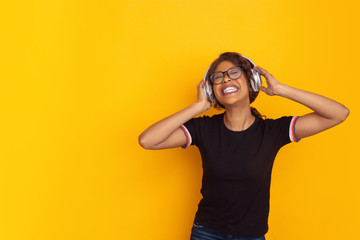 Happy listening to music. African-american young woman's portrait isolated on yellow studio background. Beautiful female curly model. Concept of human emotions, facial expression, sales, ad, youth.