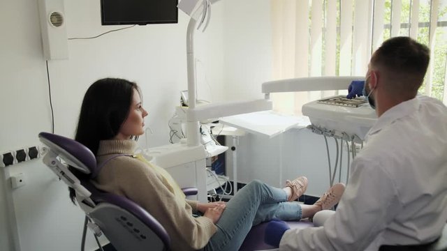 A cute girl patient looks at the dentist then leans on a dental chair and prepares for procedures.