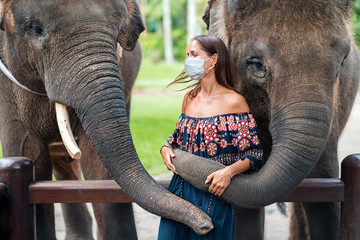 Covid-19. Portrait of a young woman in a medical mask posing with two elephants. The concept of travel during a pandemic, virus protection. Asia