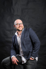 A young man in glasses and a jacket is smiling, looking at the camera and sitting on a bar stool against a black background.