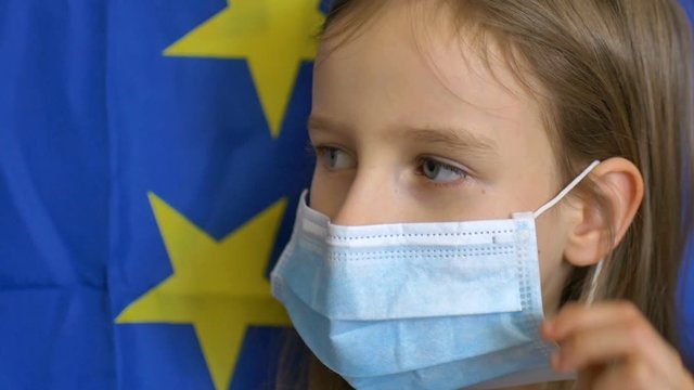 Close-up portrait of blond little girl with big blue eyes in disposible mask for protection of virus on European Union flag banner background in studio. Personal protective equipment