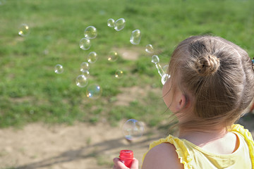 A little girl blowing soap bubbles in summer park. cute girl with space buns playing outdoors with soap bubbles. summer holiday concept. back view.