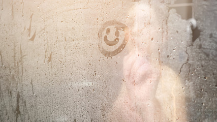 blurry naked lady draws smile on white steam condensate on glass-enclosed shower door in bathroom...