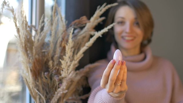 Young woman is showing yoni egg made from rose quartz stone in her hand standing near vase with spikelets indoors. Female health concept, vumfit, imbuilding or meditation