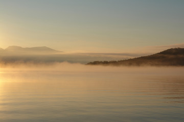 Obraz premium A scenic landscape - morning fog over the calm water of Chivyrkuisky Bay. Golden sunrise in a Zmeyevaya Bay, peninsula Svyatoy Nos (Holy Nose peninsula), Baikal Lake, Siberia, Russia