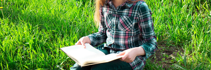 Girl teenager in a plaid shirt with long blond hair reads book outdoor sitting on green grass in park. Selective focus