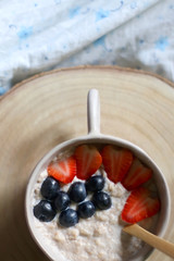 Bowl of vegan porridge with strawberry and blueberry topping, served in bed with floral sheets. Top view.
