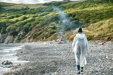 woman in a raincoat walking along a pebble beach past a sunken ship towards a beach camp. Tourism...