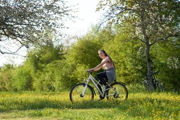 Fototapeta premium Smiling young woman on bicycle in a meadow with yellow flowers, in the background bushes and bicycles leaning on an apple tree