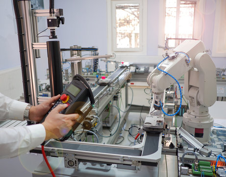 Close up of man's hand holding teach pendant to control robot arm which is integrated on smart factory production line. industry 4.0 automation line equipped with sensors and robot at background.