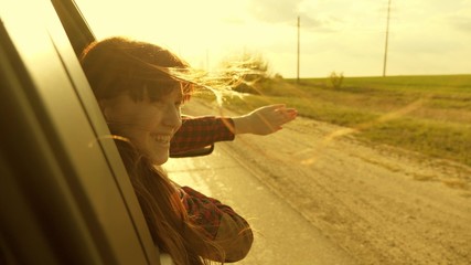 Fototapeta na wymiar free woman travels by car catches the wind with her hand from car window. Girl with long hair is sitting in front seat of car, stretching her arm out window and catching glare of setting sun