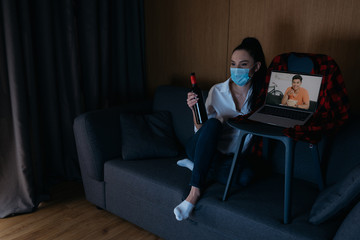 young woman in medical mask holding bottle of wine while sitting on sofa near laptop with smiling asian boyfriend on screen