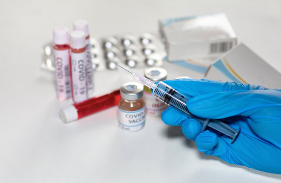 Hands Of A Doctor In Medical Gloves Holding A Hypodermic Needle Injecting Medicine From A Vaccine Bottle With View Of Medicinal Drugs And Blood Sample Vials At A Lab Shot With Selective Focus