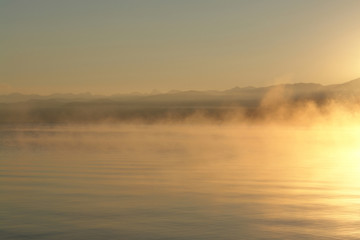 A scenic view - morning fog over the calm water of Baikal Lake. Golden sunrise at Chivyrkuisky Bay (Zabaybalsky National Park, Buryatia, Russia)