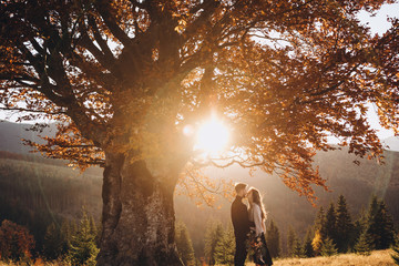 Naklejka na ściany i meble Stylish young couple in the autumn mountains. A guy and a girl stand together under a big old tree against the background of a forest and mountain peaks at sunset. The girl has a bouquet in her hands
