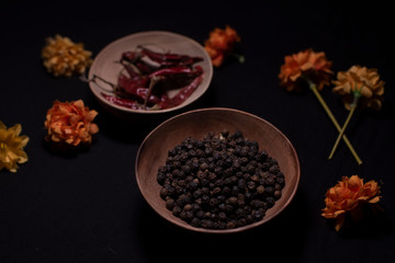 Black pepper and dried chili in a wooden bowl decorated with dry flowers in a dark copy space background.