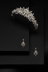 Subject shot of a silver tiara and chandelier earrings on the black stepped surface. The luxury jewelry set is adorned with moonstone gems and clear sparkling rhinestones.