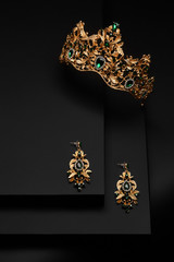 Subject shot of a golden tiara and chandelier earrings on the black stepped surface. The luxury jewelry set is adorned with emerald gems and rhinestones.