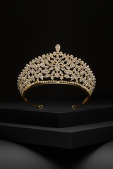 Subject shot of a golden tiara adorned with a lot of clear sparkling rhinestones. The luxury queen crown is fixed on the black stepped surface. © RedUmbrella&Donkey