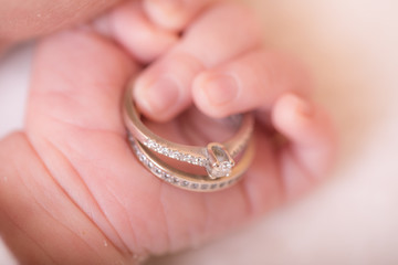 Newborn baby little hand holds parents' gold and silver wedding rings on its palm on white blanket photography.
