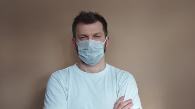 Handsome man in a white T-shirt puts on a medical protective mask and looks at the camera against a beige background. Portrait, the average plan. Natural light, soft focus.