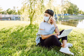 Girl working at the computer sitting on the grass near the lake in a protective medical mask on her face and have a phone talk. Freelance, quarantine concept