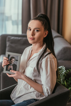 sad young woman looking at camera while holding photo of young man