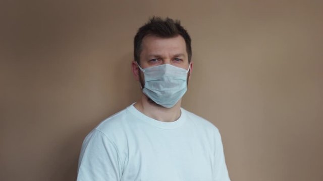 Handsome man in a white T-shirt puts on a medical protective mask and looks at the camera against a beige background. Portrait, the average plan. Natural light, soft focus.