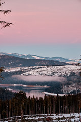 Beautiful view of lake between mountains covered with forest. Amazing sky during sunset/sunrise in winter time with a pink and blue sky.