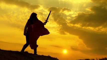 Super woman in red cloak waves a sword dreams of fighting on a mountain in the sunset light. free woman playing a superhero. girl plays roman lenin in bright rays of the sun against the sky