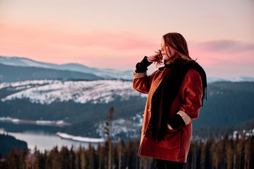 Beautiful young woman putting her hair behind her ear at golden hour with a winter lake and mountains in the background.