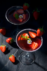 Fresh strawberry cocktail in a martini glass on dark stone background, decorated with fresh fruits and a small flower.