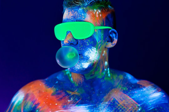 Young Man Chews Gum And Inflates A Balloon. Fluorescent Paint On Face In Studio With UV Light