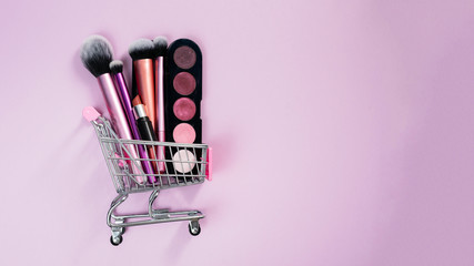 Eye shadow blush, lipstick and various makeup brushes in a pink trolley of the buyer lie on a pink...