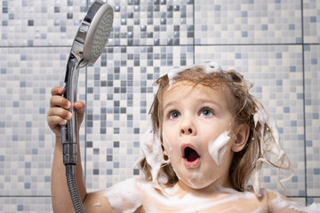 a little girl in foam is standing in the bathtub in her hand holding a shower, there is no water....