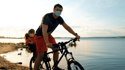 Dad driving his baby boy in protective masks on a weekend excursion with bikes on a summer day in beautiful landscape, for safety and protection they are sitting in a bike trailer