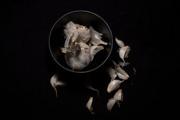 Top down image of a bowl full of garlic cloves decorated with scattered pieces in a dark copy space background. Food and product photography.