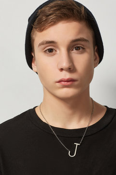 Portrait shot of a young brown-haired man with a silver necklace with a pendant in the shape of a letter J on his neck. The man is wearing in a black watch cap and a black sweatshirt. 