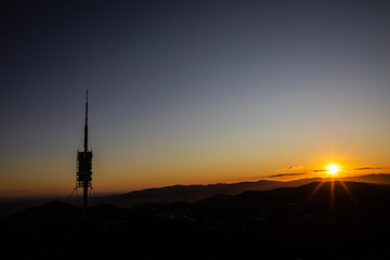 Big telecommunications antenna silhouette on the hills during sunset with sun rays