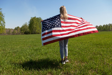Young woman with blond hair and a flag of the United States of America on her back in blue jeans and sneakers in a field against a background of green trees and grass.