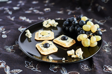 Three blue cheese crackers with grapes are on a black plate.