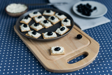 Plate with crackers with soft cheese and olives on a blue polka dot tablecloth.