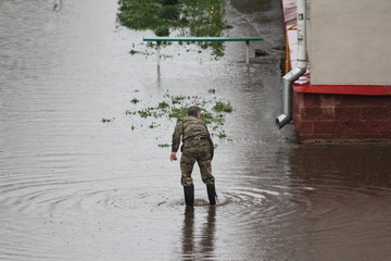 A service man in camouflage clothing searches for a sewer grate under water in a large puddle against the background of a drainpipe at home, flooding after rain in the city