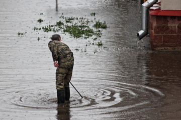 A service man in camouflage clothing searches for a manhole under water in a large puddle against the background of a drainpipe at home, flooding after rain in the city