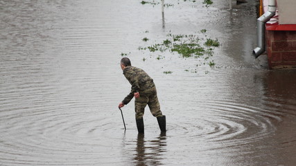 A service man in camouflage clothing searches for a gutter hatch under water in a large puddle against the background of a drainpipe at home, deluge after rain in the city