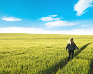 Young blonde girl running through the green wheat field and blue sky with  white clouds. Concept of travel, inspiration, freedom, lifestyle with copy space.