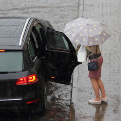 A young woman with an umbrella stands on the sidewalk at the open rear right door of a black SUV on a road in the city on a summer day in a deep puddle after rain
