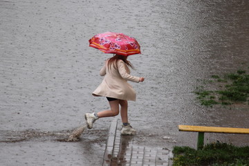 One girl in a short light coat under a colored umbrella runs through a huge puddle in the Park...