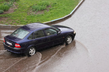 Fototapeta premium A dark blue car drives into a large puddle on the background of a curb and green grass, heavy rain in the city