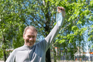 Happy middle-aged gray-haired man taking off a medical mask from his face in the spring park. Over coronavirus. End of pandemic,  epidemic and quarantine. Concept